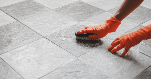 Person cleaning floor tiles with a brush and orange rubber gloves
